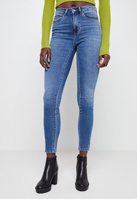 Jeans ajustados de mezclilla azul con cintura de altura media, ligero desgaste y costuras en contraste; combinados con botas de tacón negro hasta el tobillo.