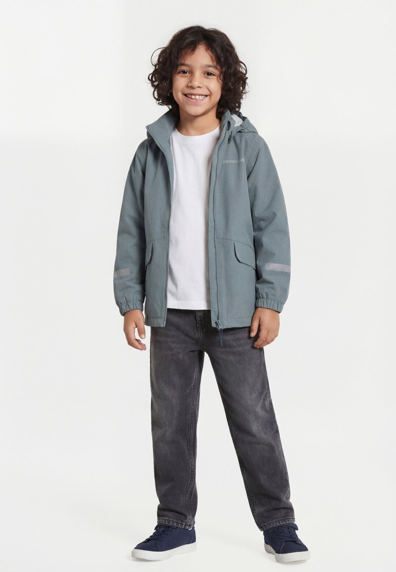 Παιδί με σγουρά μαλλιά που χαμογελά, φοράει γαλάζιο μπουφάν, λευκό μπλουζάκι, γκρι τζιν και ναυτικά αθλητικά παπούτσια, στέκεται μπροστά σε λευκό φόντο.