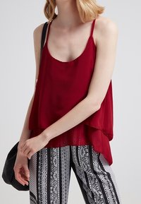 Burgunderfarbene ärmellose Bluse mit verstellbaren Trägern, lockerer Passform und geschichtetem Saum. Kombiniert mit Hose im schwarz-weißen Streifenmuster.