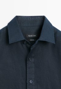 Tmavě modrá lněná košile s knoflíky a špičatým límečkem, s viditelnou značkou "Massimo Dutti" a štítkem "100 % len, vyrobeno v Číně" uvnitř límečku.