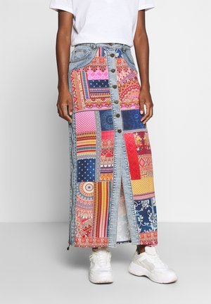 Personne portant une jupe en jean patchwork multicolore boutonnée, des baskets blanches et un haut blanc uni à manches courtes, debout devant un fond uni.