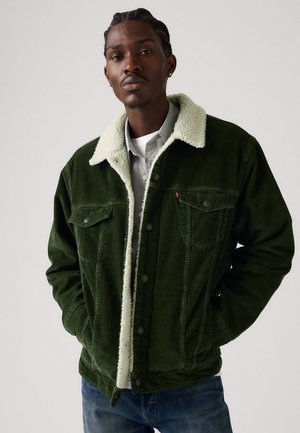 Groene corduroy jas met een witte nep-shearling kraag, voorzakken en drukknoopjes. Draag het over een lichtgrijs shirt.