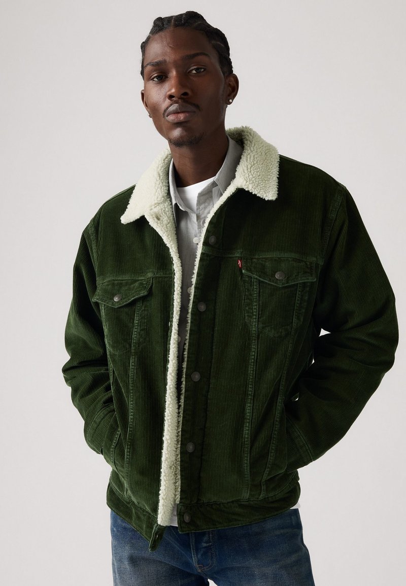 Green corduroy jacket with a white faux shearling collar, front pockets, and snap buttons. Worn over a light gray shirt.
