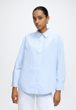 Femme aux cheveux foncés portant des boucles d'oreilles créoles dorées et une chemise boutonnée à rayures bleu clair avec un pantalon blanc sur un fond uni.