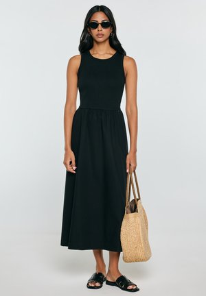 Kobieta w czarnej sukience midi bez rękawów, czarne sandały typu slide, okulary przeciwsłoneczne, trzyma plecioną słomianą torebkę na jednolitym tle.