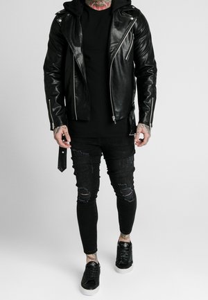 Mann trägt eine schwarze Leder-Bikerjacke, zerrissene schwarze Skinny-Jeans, schwarze Turnschuhe und sichtbare Tätowierungen am Hals und an den Händen.