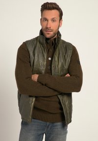 Olivgrüne Lederweste über einem braunen Strickpullover, ausgestattet mit einem hohen Kragen und Knopfdetails, getragen mit blauen Jeans.