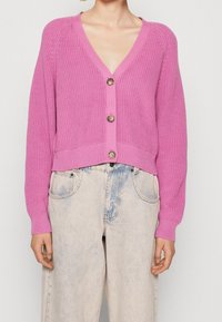 Różowy dziergany kardigan z dekoltem w serek, trzema guzikami i prążkowanym dołem, w połączeniu z jasnymi dżinsowymi spodniami. Teksturowana tkanina z długimi rękawami.