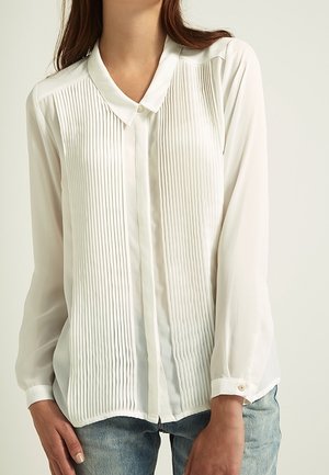 Femme portant un chemisier blanc à manches longues avec des plis verticaux sur le devant, des poignets boutonnés, et un jean bleu clair.