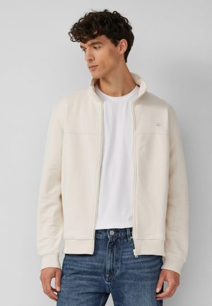 Lichtbeige zip-up jack met een hoge kraag, gemaakt van zachte stof. Heeft een effen ontwerp en een klein logo op de borst. Gecombineerd met een wit T-shirt.