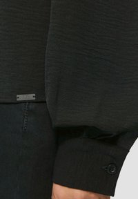 Czarna, długa bluzka z długim rękawem, uszyta z teksturowanej tkaniny, z zapinanym mankietem i małą metką na dolnym brzegu. Prosty i elegancki design.