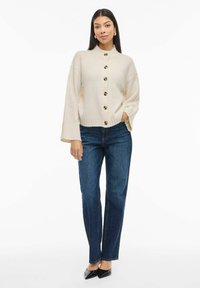 Elfenbensfarget strikket cardigan med knappestengning og ribbestrikket kant, kombinert med mørkeblå jeans med rette ben og svarte spisse hæler.