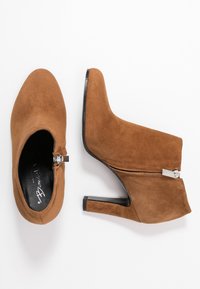 Brune ruskindsankelstøvler med spidse tåform, lynlåslukning og slanke sorte hæle. Glat tekstur og minimalistisk design, velegnet til formel påklædning.