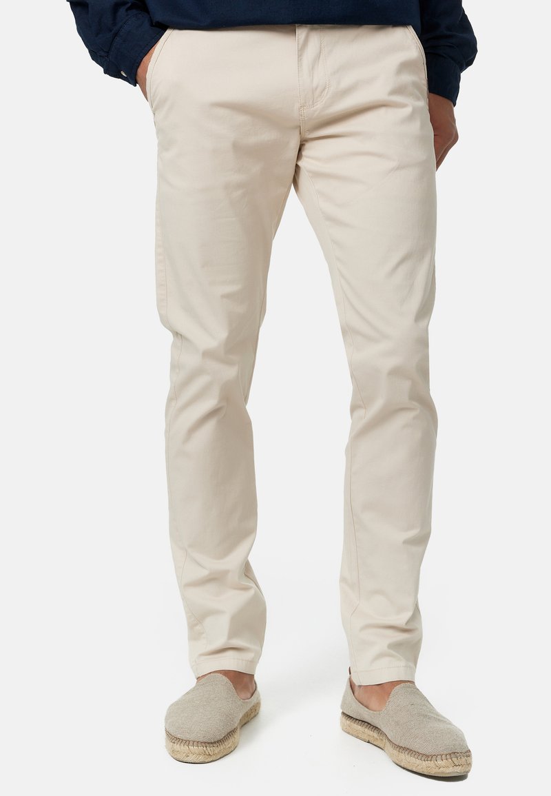 Pantalon en coton beige à coupe droite, doté de poches latérales, de légères plis et associé à des espadrilles gris clair.
