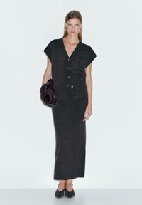 Donna che indossa un cardigan senza maniche grigio scuro con bottoni, gonna abbinata alla caviglia, scarpe nere e tiene in mano una borsa di pelle scura.