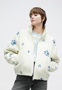 Fleecejakke med glidelås i kremfarge med blå blomsterbroderi. Har høy krage, elastiske mansjetter og myk tekstur, brukt over en lyseblå topp med knapper.