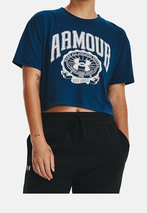 Osoba nosząca niebieską krótka koszulkę Under Armour z nadrukowanym logo i czarne spodnie ze sznurkiem, ręka oparta przy boku.