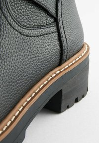 Botte en cuir noir texturé avec un accent marron sur la semelle, présentant un talon épais et un détail de couture blanche le long du bord.