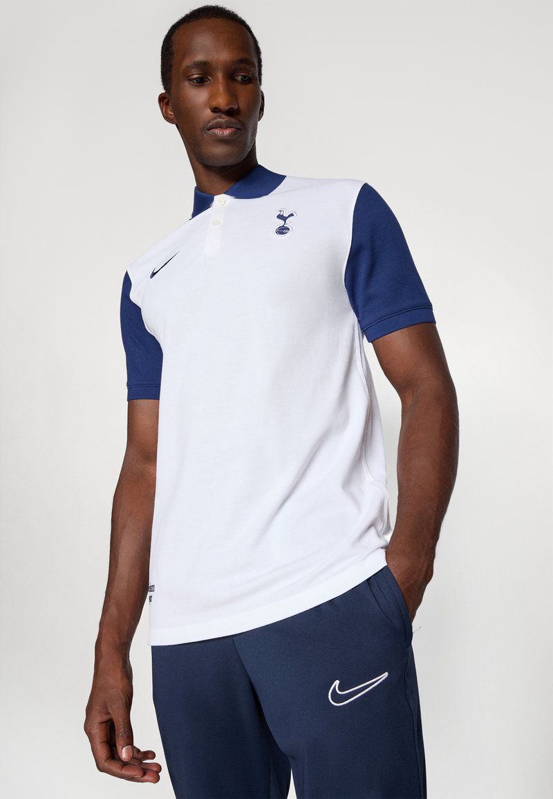 Polo-shirt blanc et bleu marine avec un col, marqué d'un logo bleu marine. Matériau en coton, manches courtes, complété par un pantalon bleu marine avec un swoosh blanc.