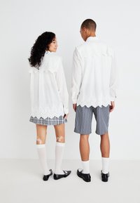 Hvit bluse med brodert blondekant, kombinert med et rutete skjørt. Modellen har på seg stripete shorts, knelange sokker og svarte sko.