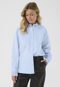 Light blue button-up shirt with a collar, long sleeves, and eyelet detailing on the cuffs. Paired with black leather trousers.