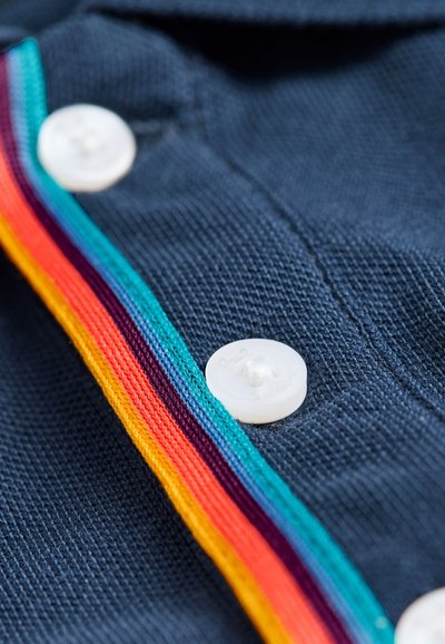 Tengerészkék szövet texturált kötött felülettel, fehér gombbal és csíkozott szalaggal, amely piros, narancssárga, sárga, tengerzöld és lila színekben pompázik.