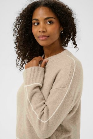 Béžový sveter s mäkkou textúrou, rebrovanými manžetami a bielymi detailmi, s voľným strihom a okrúhlym výstrihom.