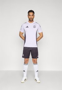 Futbalový dres s krátkym svetlofialovým rukávom s čiernymi prvkami, čierne nohavice, biele ponožky a kopačky. Zobrazené logo Manchester United.