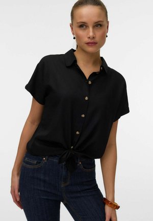 Kvinne med svart knappeskjorte med korte ermer, knytt i livet, mørkeblå jeans, sorte øredobber og et ravarmbånd på høyre håndledd.