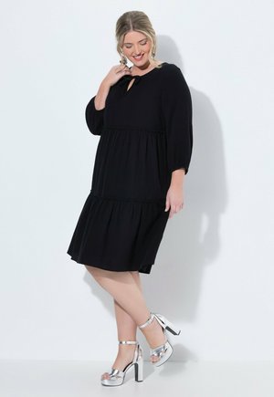 Frau steht vor einer weißen Wand und trägt ein lockeres schwarzes Kleid mit Dreiviertelärmeln und silberne High-Heel-Sandalen.