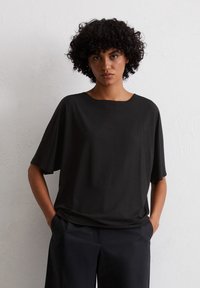 Czarny krótki top z miękkiego materiału, z okrągłym dekoltem i luźnym krojem, w połączeniu z czarnymi spodniami na neutralnym tle.