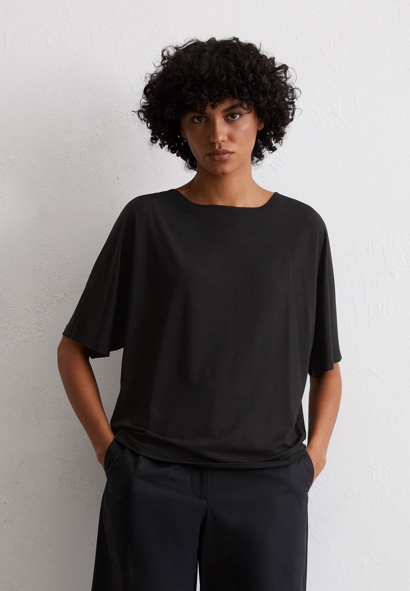 Czarny krótki top z miękkiego materiału, z okrągłym dekoltem i luźnym krojem, w połączeniu z czarnymi spodniami na neutralnym tle.