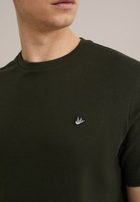 Zeleno bombažno majico s krožnim vratom, ki ima na prsih majhno belo vezenje ptička, gladko teksturo in prilegajoč dizajn.