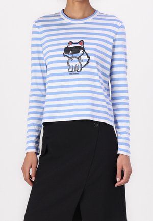 Femme portant un chemisier à manches longues rayé bleu et blanc avec un chat cartoon en lunettes de soleil sur le devant et une jupe noire.