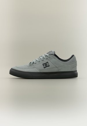 Baskets basses gris DC avec semelle en caoutchouc noir et lacets, avec des perforations sur l'avant-pied et le logo DC sur le côté.
