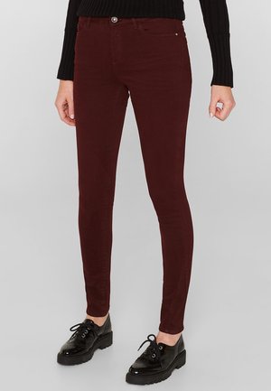 Persoon die donkerrode slim-fit broek en zwarte lakleren veterschoenen draagt, staand tegen een lichtgrijze achtergrond.