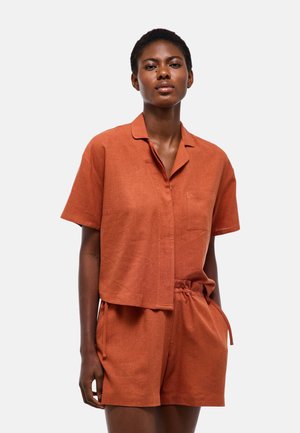 Femme portant une chemise rouille-orange à manches courtes avec boutons et un short assorti à cordon, debout devant un fond clair uni.