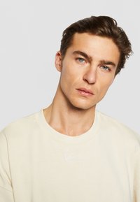 Kremfarget sweatshirt med rund hals, korte ermer og brodert logo i hvitt på brystet; myk stoffkvalitet og avslappet passform.