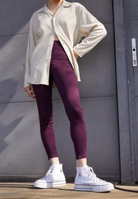 Camisa de manga larga color beige claro, mallas moradas y zapatillas altas blancas. La camisa tiene cuello y botones, mientras que las mallas son ajustadas.