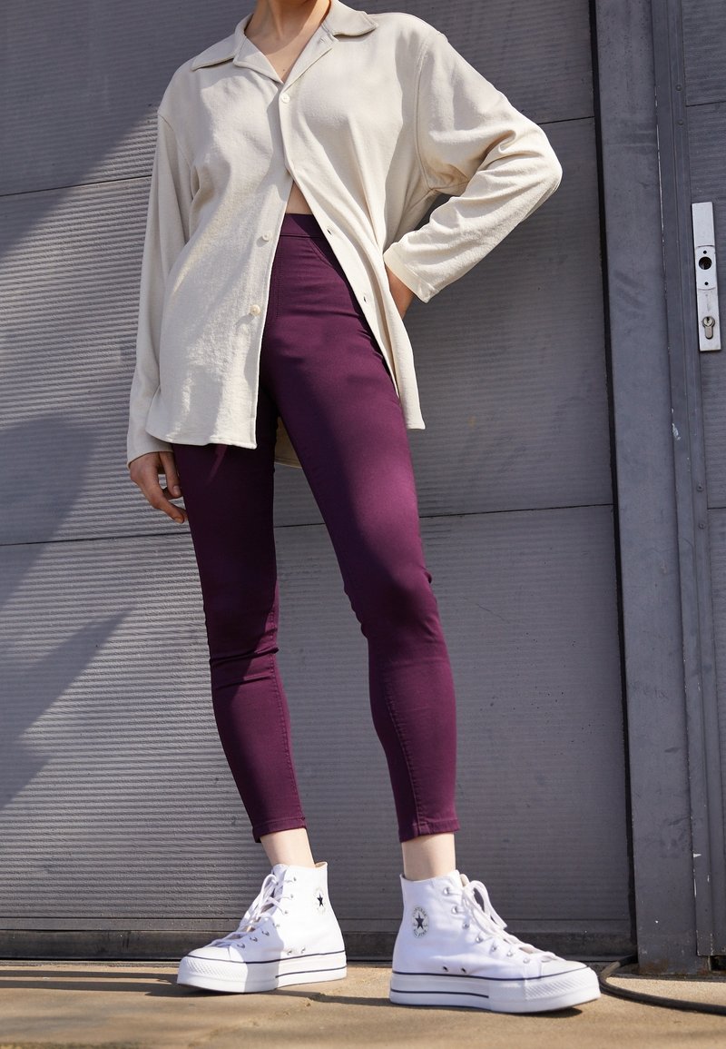 Camisa de manga larga color beige claro, mallas moradas y zapatillas altas blancas. La camisa tiene cuello y botones, mientras que las mallas son ajustadas.