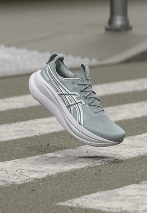 Pantof de alergare albastru deschis cu talpă albă și logo-ul Asics, în aer deasupra unei treceri de pietoni pe un drum asfaltat.