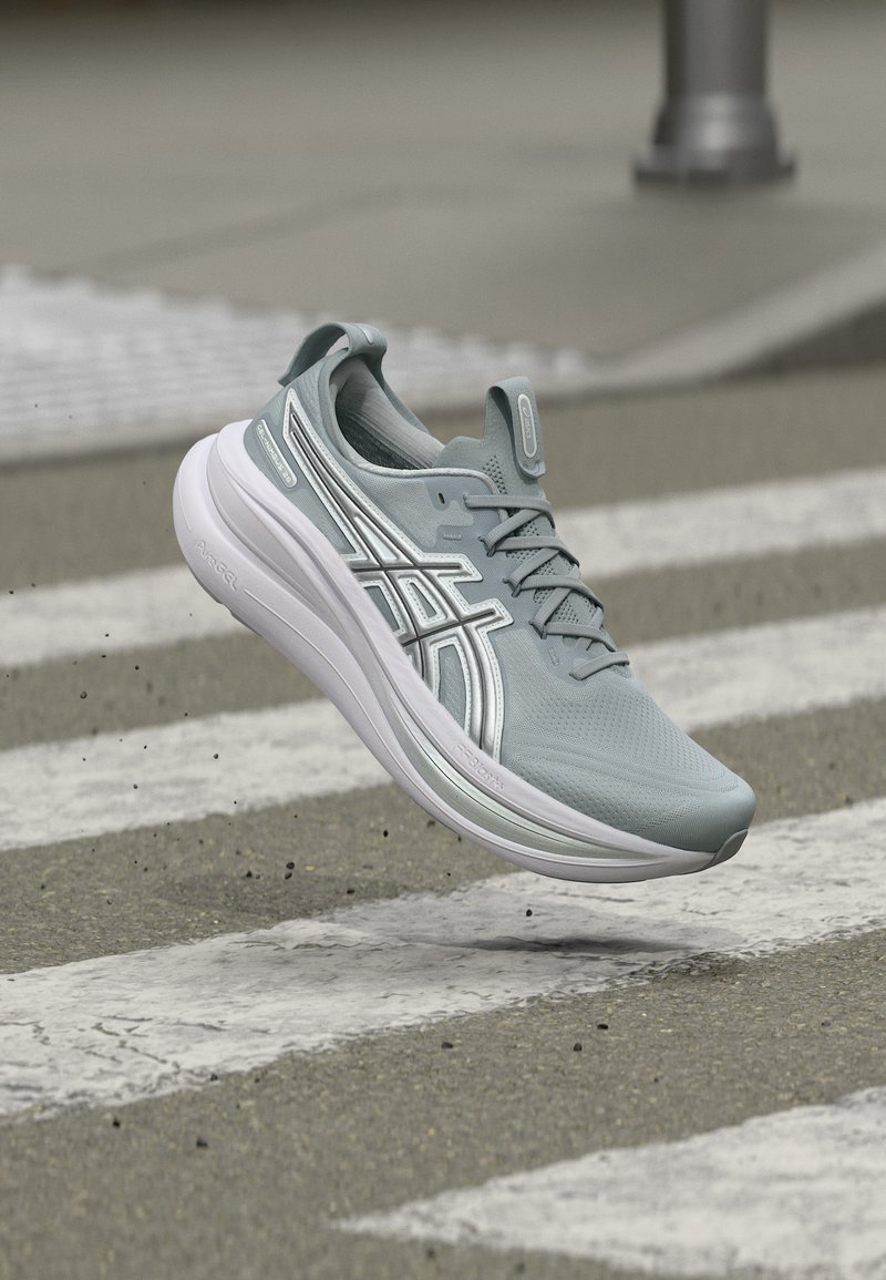 Ανοιχτό μπλε αθλητικό παπούτσι με λευκή σόλα και λογότυπο Asics, στον αέρα πάνω από διάβαση πεζών σε ασφάλτινο δρόμο.