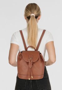 Sac à dos en cuir marron avec une finition texturée, fermeture à rabat rond, bretelles ajustables et quincaillerie dorée, vu de l'arrière.