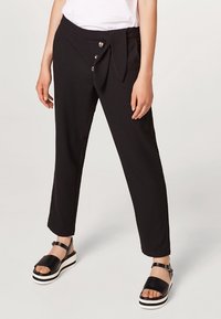 Pantaloni neri con dettaglio in vita annodato, caratterizzati da una texture liscia e tre bottoni decorativi sul davanti. Abbinati a sandali con zeppa.