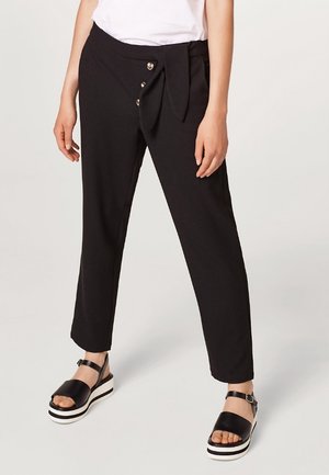 Pantaloni neri con dettaglio in vita annodato, caratterizzati da una texture liscia e tre bottoni decorativi sul davanti. Abbinati a sandali con zeppa.