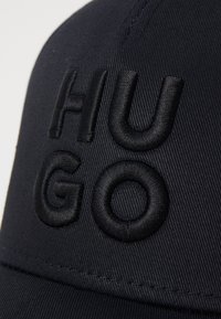 Căciulă bleu marin dintr-un material texturat, cu litere negre brodate în relief "HUGO" pe partea din față, având un vizor curbat și cusături detaliate.
