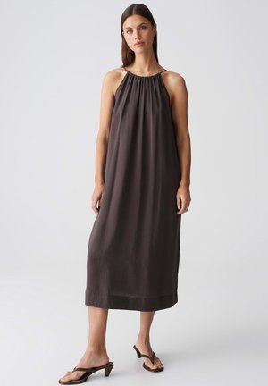 Frau mit glatten braunen Haaren steht und trägt ein ärmelloses dunkelbraunes Midikleid und braune Sandaletten mit Absatz vor einem schlichten hellen Hintergrund.