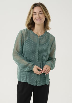 Groene doorzichtige blouse met lange mouwen, knoopsluiting, geplooide textuur en een klein stippenpatroon. Draagt over zwarte broek.