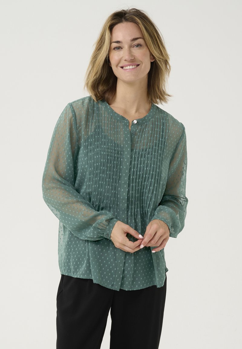 Blusa trasparente verde con maniche lunghe, chiusura con bottoni, texture plissè e piccolo motivo a pois. Indossata sopra pantaloni neri.
