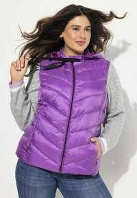 Gilet imbottito viola con design a zigzag, chiusura con zip e cappuccio regolabile. Abbinato a un maglione grigio chiaro e jeans scuri.